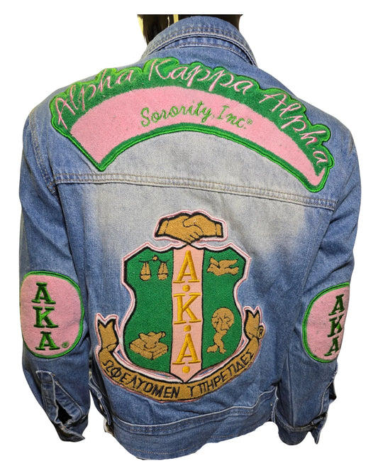 View of the back of a light-wash Alpha Kappa Alpha AKA denim jacket. The back features a large green chenille patch in the shape of the AKA shield/crest, surrounded by various smaller pink and green patches.