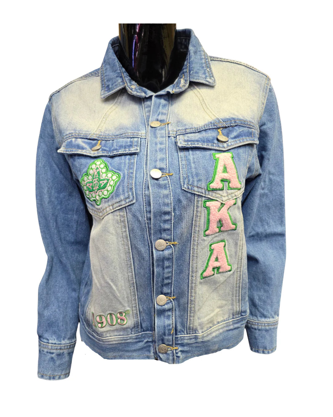 Close-up of the front of a light-wash Alpha Kappa Alpha AKA denim jacket. The jacket features pink and green chenille patches with the letters AKA, a large green ivy leaf patch, and a small patch on the bottom left reading "1908."