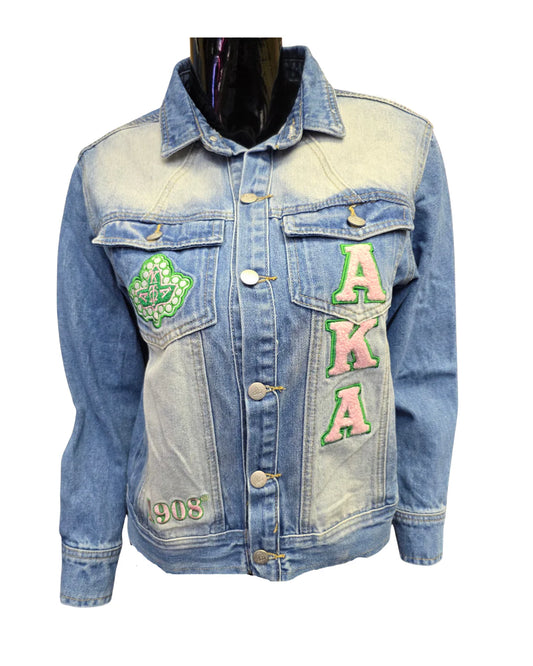 Close-up of the front of a light-wash Alpha Kappa Alpha AKA denim jacket. The jacket features pink and green chenille patches with the letters AKA, a large green ivy leaf patch, and a small patch on the bottom left reading "1908."