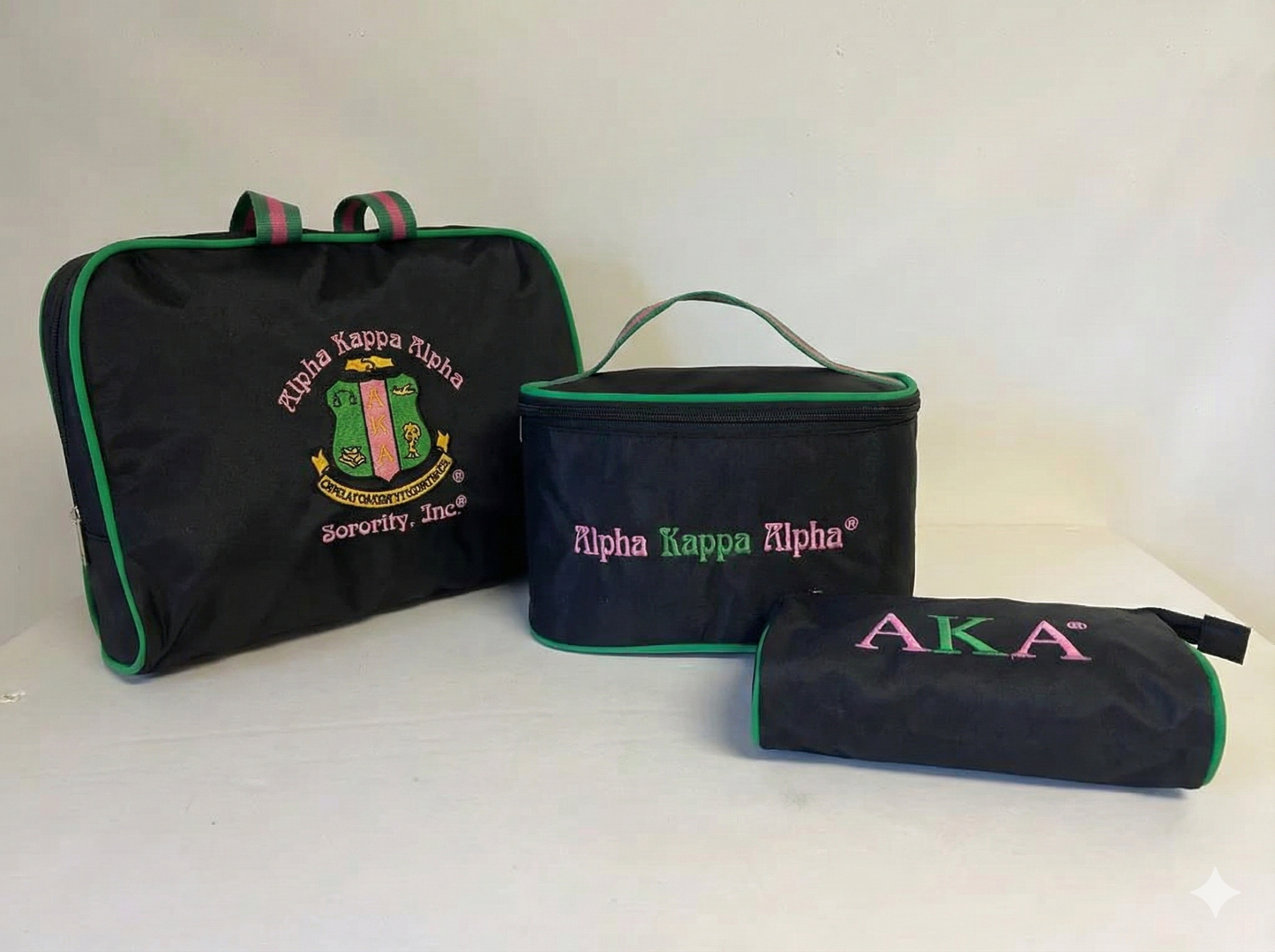 A three-piece black travel cosmetic bag set arranged on a white background. The set features pink and green accents and embroidery representing the Alpha Kappa Alpha sorority. It includes a large rectangular toiletry bag with handles featuring the full sorority crest and name, a medium boxy makeup case with "Alpha Kappa Alpha" text, and a small zipper pouch with "AKA" letters.