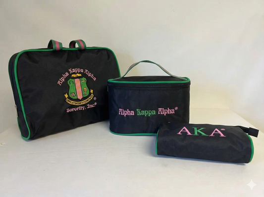 A three-piece black travel cosmetic bag set arranged on a white background. The set features pink and green accents and embroidery representing the Alpha Kappa Alpha sorority. It includes a large rectangular toiletry bag with handles featuring the full sorority crest and name, a medium boxy makeup case with "Alpha Kappa Alpha" text, and a small zipper pouch with "AKA" letters.