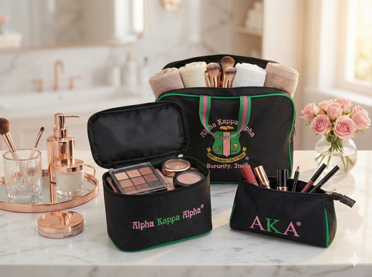 A three-piece black cosmetic bag set representing Alpha Kappa Alpha sorority. The set includes a large rectangular toiletry bag with the full sorority crest and handles, a medium boxy makeup case with "Alpha Kappa Alpha" script, and a small zipper pouch with "AKA" letters. All bags feature signature pink and green embroidery and trim on a black background.