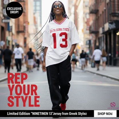 : African American woman wearing a white Delta Sigma Theta sorority jersey tee with "DELTA SIGMA THETA" and "13" in red on the front, paired with black baggy pants, red sneakers, and sunglasses, walking on a city street with graffiti walls.