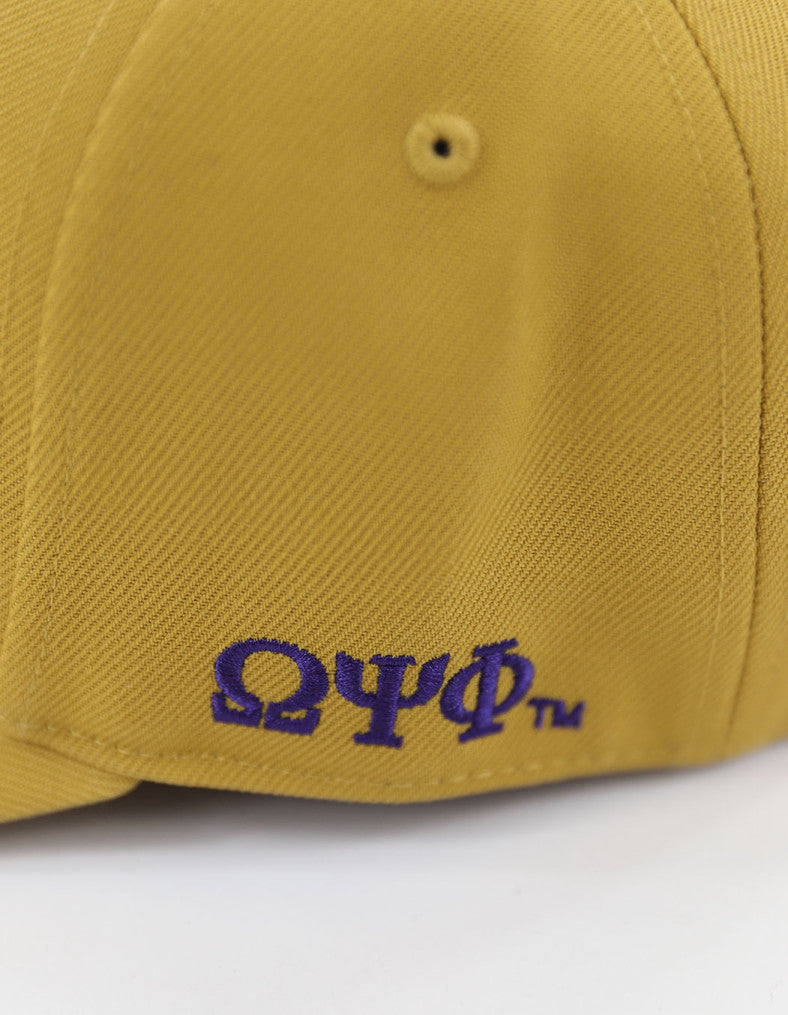 Close-up detail of purple Omega Psi Phi Greek letters embroidered on the side of a textured old gold baseball cap.