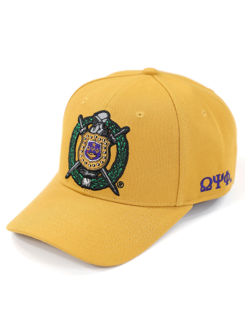 Front and side view of an Old Gold Omega Psi Phi fraternity cap featuring a large, detailed embroidered crest shield.